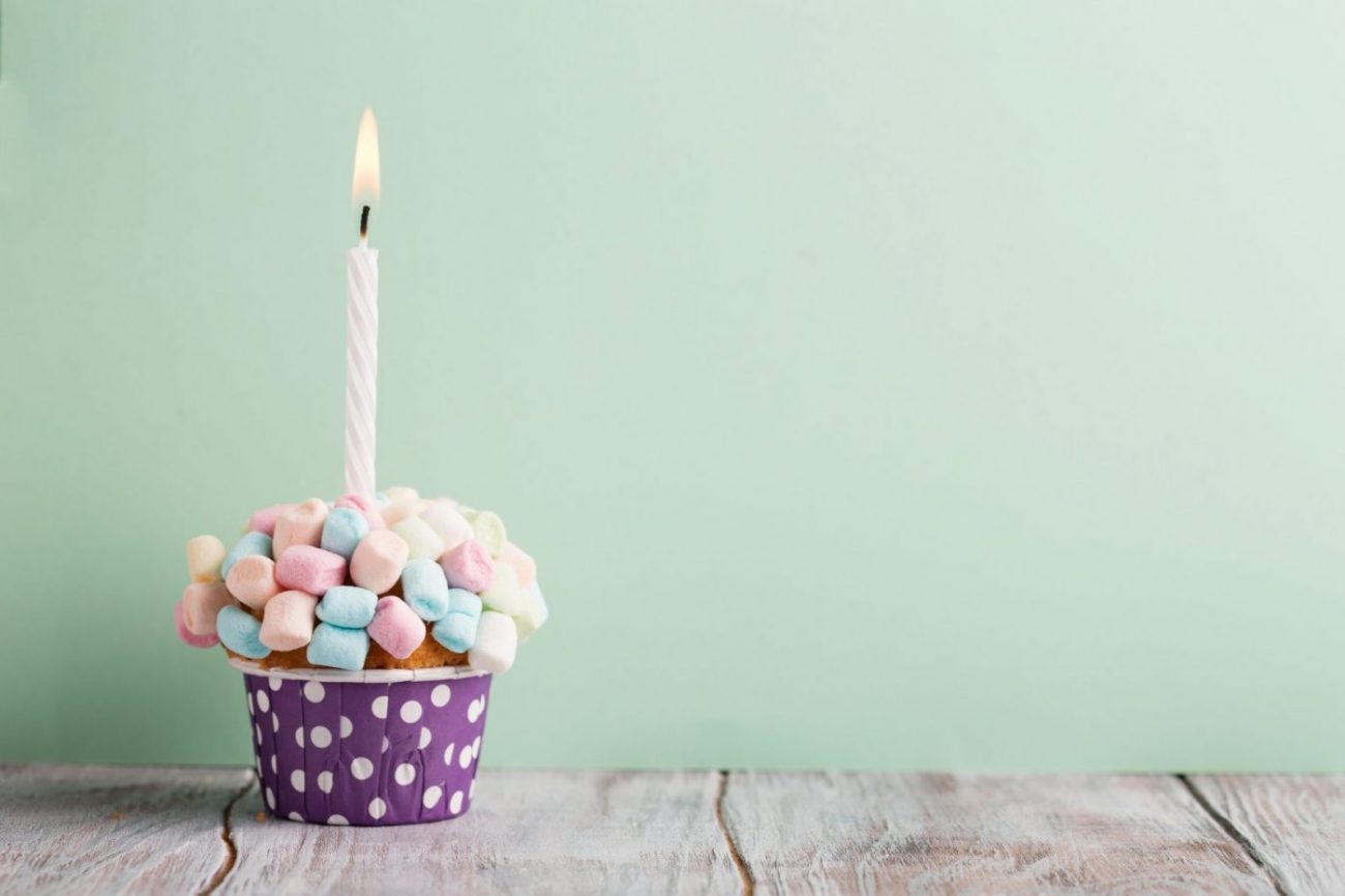 Delicious birthday vanilla cupcake with colorful marshmallows, cream and lighted candle on a green mint background, selective focus.
